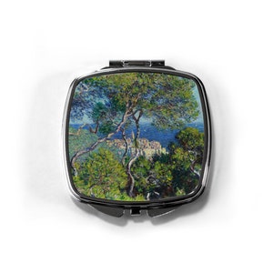 May include: A silver compact mirror with a colorful image of a landscape painting. The painting features a green hillside with a town in the distance and a blue sky with white clouds.