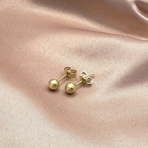 May include: A pair of gold-toned stud earrings with a textured, round design. The earrings are displayed on a soft, pink fabric background.