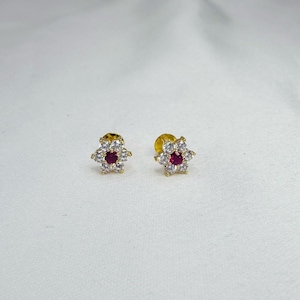 Op de afbeelding: Een paar goudkleurige oorstekers. Elke oorbel heeft een bloemdesign met een centrale rode edelsteen omringd door witte edelstenen. De oorbellen zijn geplaatst tegen een witte achtergrond.