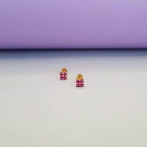 Pode incluir: Um par de brincos dourados, cada um com um grupo de pequenas pedras preciosas rosa brilhantes dispostas em forma quadrada. Os brincos estão sobre um fundo branco e roxo.