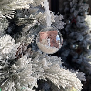May include: A clear glass Christmas ornament filled with white material, a small cup, and a packet with the word "Chipotle" printed on it. The ornament hangs from a white ribbon on a flocked Christmas tree.