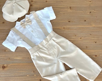 May include: A beige outfit for a baby or toddler, including a white shirt with suspenders, beige pants, and a beige newsboy cap with a bow tie.