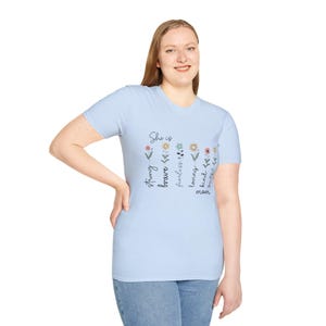 Op de afbeelding: Lichtblauw T-shirt met de tekst "She is" boven een rij kleurrijke bloemen en de woorden "strong, brave, fearless, loving, kind, mom" in een verticale lettertype. Het shirt is gemaakt van een zacht materiaal.