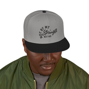 Puede incluir: Gorra de béisbol gris y negra con el texto "NOT BY MY Strength BUT HIS" en escritura negra. La gorra tiene una visera plana y un botón en la parte superior. La persona de la imagen lleva una chaqueta verde.