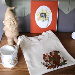 May include: A cream-coloured t-shirt featuring an embroidered squirrel holding an acorn surrounded by autumn leaves. The shirt is displayed near a ceramic garden gnome, a white floral watering can, and a framed sunflower illustration on an orange background.