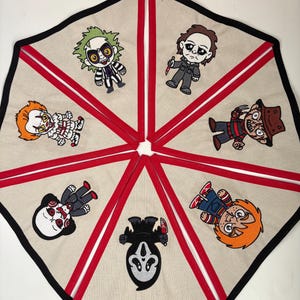 May include: A beige tree skirt with embroidered cartoon horror movie characters. The skirt has red ribbon accents and a black border. Characters include Beetlejuice, Michael Myers, Freddy Krueger, and Chucky.