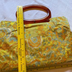 May include: A vintage yellow, orange and blue floral patterned handbag with a brown plastic top handle. The handle is shaped like an oval. The bag is made of fabric and has a quilted texture.