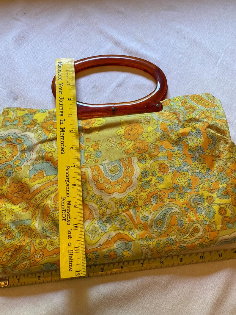 May include: A vintage yellow, orange and blue floral patterned handbag with a brown plastic top handle. The handle is shaped like an oval. The bag is made of fabric and has a quilted texture.