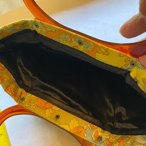 May include: A vintage handbag with a floral fabric exterior and a black satin lining. The bag has a brown plastic handle with a unique double loop design.