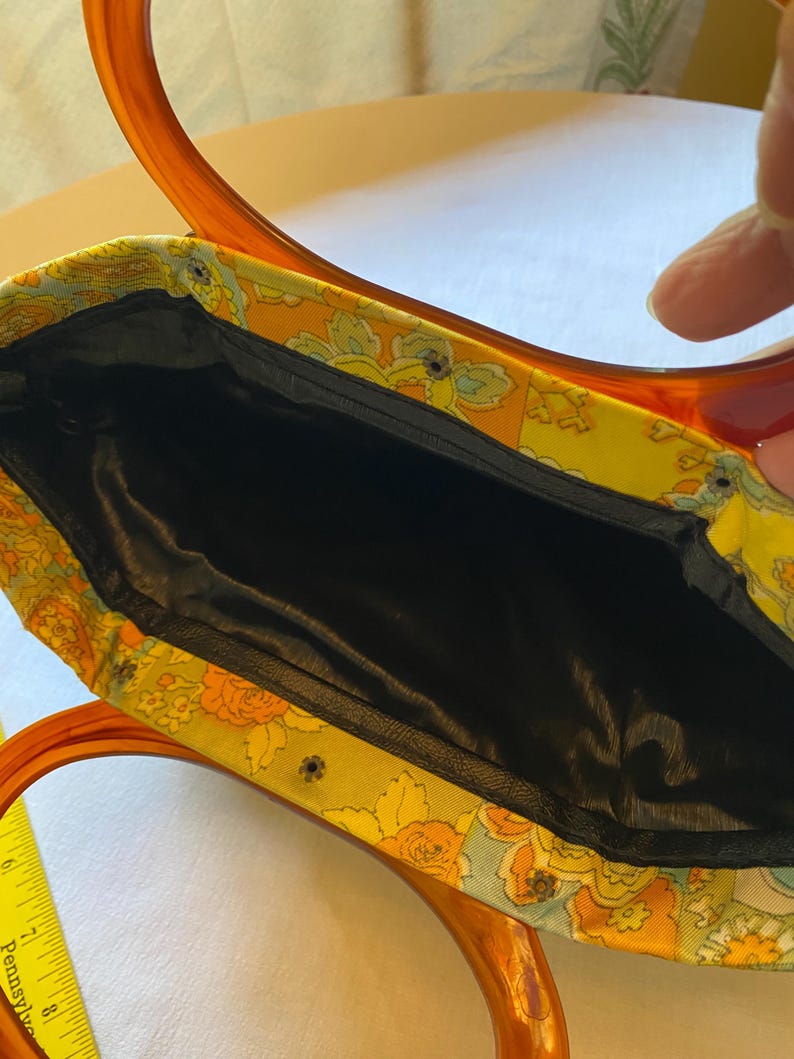 May include: A vintage handbag with a floral fabric exterior and a black satin lining. The bag has a brown plastic handle with a unique double loop design.