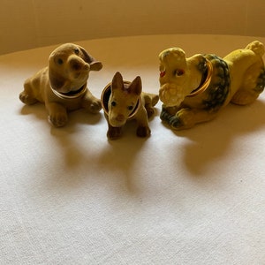 May include: Three small, plush dog figurines with gold collars. The dogs are brown and tan with black accents. The dogs are sitting on a white surface.
