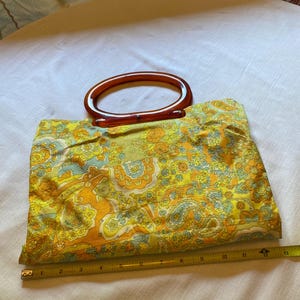 May include: A vintage yellow, orange and blue paisley patterned fabric handbag with a brown plastic handle.