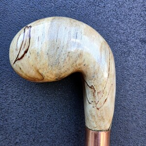 May include: A wooden walking stick with a curved handle. The handle is light-coloured with dark brown streaks and a copper-coloured metal band. The lower portion of the stick is a darker brown colour.