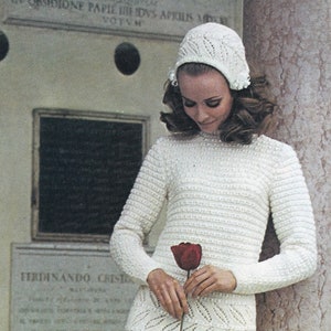 Puede incluir: Un vestido blanco de ganchillo con un patrón de hojas y un sombrero blanco de ganchillo a juego. El vestido es largo y tiene un cuello alto. La modelo sostiene una sola rosa roja.