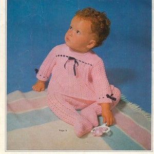 May include: A baby wearing a pink knitted outfit with black trim and a black ribbon. The baby is sitting on a striped blanket and looking up. The image is from a knitting pattern book titled "26 New Designs for Baby Knitteds" by Patons, Book 972, Page 9.