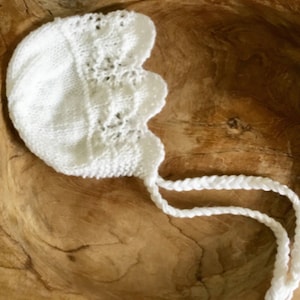 May include: White crocheted baby bonnet with a scalloped edge and long ties. The bonnet is lying in a wooden bowl.