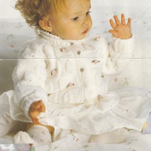 Op de afbeelding: Een wit gebreid babyvest met knoopsluiting en kleine roze bloemetjes.