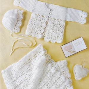 Könnte beinhalten: Ein weißes Häkelset für Babys bestehend aus Mütze, Jacke und Decke. Das Set ist mit einer zarten, durchbrochenen Masche gearbeitet.