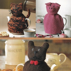 Könnte beinhalten: Drei gestrickte Teekannenwärmer in Form einer Katze, eines Schweins und eines Bären. Die Katze ist schwarz und orange, das Schwein ist rosa und der Bär ist braun. Die Wärmer sitzen alle auf Teekannen.