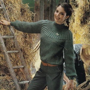 Könnte beinhalten: Ein grüner Strickpullover mit hohem Kragen und einem braunen Gürtel. Der Pullover hat eine gerippte Struktur und einen leicht bauschigen Ärmel. Das Model trägt den Pullover mit einer dunklen Hose und steht in einer Scheunenkulisse.