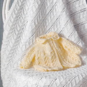 May include: A white crocheted baby blanket with a delicate diamond pattern is draped over a white wicker chair. A small, pale yellow crocheted baby jacket is sitting on the blanket.