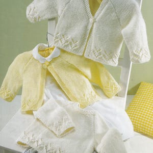 Könnte beinhalten: Drei gestrickte Baby-Cardigans in Creme und Gelb werden auf einem weißen Stuhl präsentiert. Die Cardigans haben dekorative Kanten und sind aus weichem Garn gefertigt. Ein gelber Cardigan hat einen weißen Kragen. Die Cardigans sind im klassischen Stil.