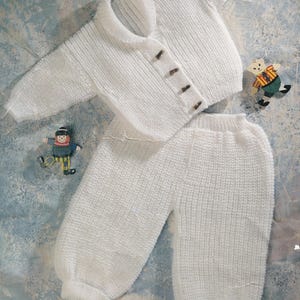 May include: A white knitted baby outfit with a cardigan and trousers. The cardigan has a collar and buttons.