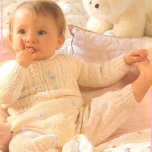 May include: A baby wearing a cream-colored knitted outfit, including a sweater and pants, with delicate floral accents. The image features the text "Baby Book" at the top and "18 smart knits for small people" at the bottom. A plush polar bear is in the background.