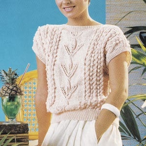 May include: A pale pink knitted top with short sleeves and a round neckline. The top has a central leaf pattern and cable knit detailing. The model is wearing a white bracelet and white skirt. The background is a blue wall with a pineapple drink.