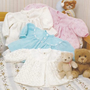 Puede incluir: Cuatro jerséis de punto para bebé en blanco, rosa, azul y blanco con un patrón de encaje. Los jerséis son abotonados y tienen un dobladillo con volantes.