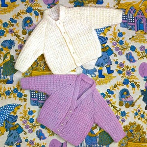 Könnte beinhalten: Zwei gestrickte Baby-Cardigans, einer weiß und einer lila, mit Knopfverschlüssen. Die Cardigans liegen auf einem floralen Stoffhintergrund.