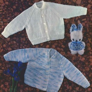 May include: Two knitted baby cardigans are shown, one plain white and the other blue and white striped, both with button fastenings. A small stuffed bunny with blue detailing is also present. The items are on a brown, textured surface.