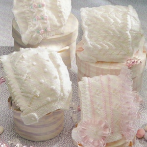 以下が含まれることがあります： 薄い紫色の背景に、異なるパターンと装飾が施された白いニットのベビーボンネットが4つ、箱の上に置かれています。ボンネットは繊細なレースとパールがあしらわれています。