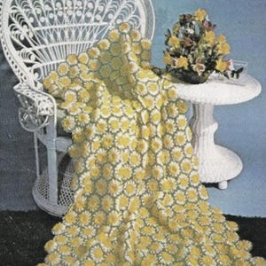 May include: A crochet blanket featuring a repeating pattern of yellow and white daisy-like floral motifs connected by green yarn. The blanket is draped over a white wicker chair, showcasing a textured, handmade construction with a scalloped edge.