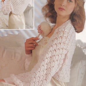 May include: A white, open-weave crochet shrug with long sleeves. The bolero-style top features a delicate, lacy pattern and a rounded neckline. The sleeves have a slight bell shape. The garment is shown over a satin dress.