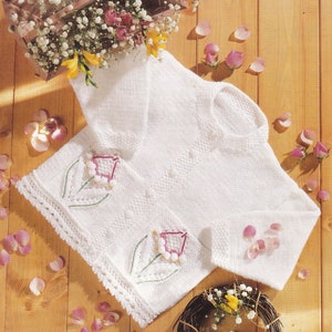 May include: White knitted baby cardigan with embroidered flowers. The cardigan has a button closure and a delicate lace trim.