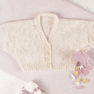 May include: Cream-colored, hand-knitted baby cardigan with a V-neck and button closure. The sweater is made of soft yarn and has a delicate texture. The garment is displayed on a pink patterned surface.