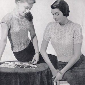 May include: Two women wearing white knitted sweaters with a lacy pattern. The women are sitting at a table with cards spread out on the surface.
