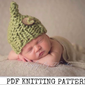 Peut inclure: Un bonnet en tricot vert pour bébé avec un bouton en bois. Le bonnet a un sommet pointu et est fait avec un fil épais. Le bonnet est sur un bébé endormi sur une couverture de couleur crème.