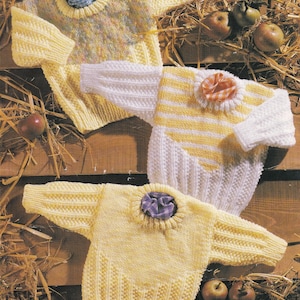 May include: Three knitted baby sweaters in yellow, white, and a multi-colored pattern. The sweaters are laid out on a rustic wooden surface with straw and apples in the background.