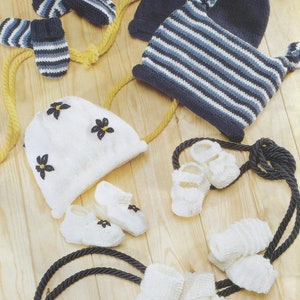 Puede incluir: Un conjunto de artículos de punto para bebé que incluye un gorro blanco con adornos de flores negras, un gorro a rayas azul y blanco con orejeras, un par de botines blancos y dos pares de calcetines blancos.