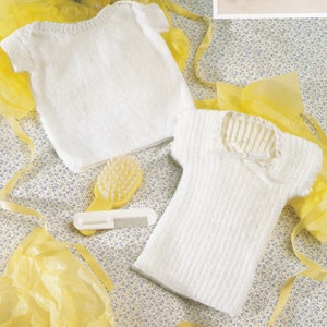Pode incluir: Duas camisas de bebé de malha branca, uma de manga curta e a outra de manga comprida, sobre um fundo estampado amarelo e branco.
