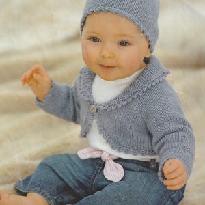 May include: A baby wearing a light blue knitted cardigan with a button closure and a matching light blue knitted hat. The cardigan has a crocheted trim around the neckline and the hat has a crocheted edge. The baby is also wearing blue jeans and a white shirt with a pink and white gingham ribbon tied around the waist.
