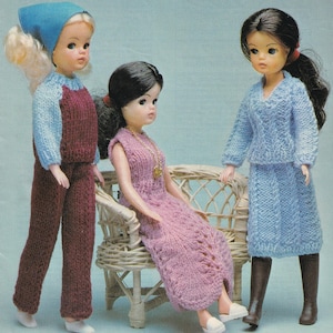 Könnte beinhalten: Drei Puppen in gestrickten Outfits. Eine Puppe trägt ein blaues und weinrotes Outfit mit einem blauen Kopftuch. Eine andere Puppe trägt ein rosa Kleid und eine goldene Halskette. Die dritte Puppe trägt einen blauen Pullover und Rock mit braunen Stiefeln.
