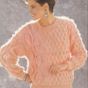 Könnte beinhalten: Ein hellpfirsichfarbener Pullover mit Rundhalsausschnitt und langen Ärmeln. Der Pullover hat ein strukturiertes Muster auf der rechten Seite und ein Blattdesign auf der linken Seite. Die Bündchen und der Saum sind gerippt.