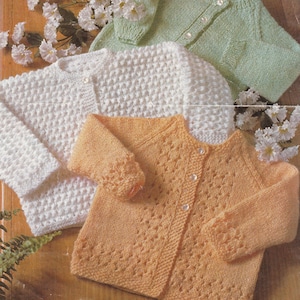 Może przedstawiać: Trzy dzianinowe kardigany dla niemowląt w kolorze białym, pomarańczowym i zielonym. Biały kardigan ma fakturowany wzór splotu, pomarańczowy kardigan ma ażurowy wzór splotu, a zielony kardigan ma prosty wzór splotu.
