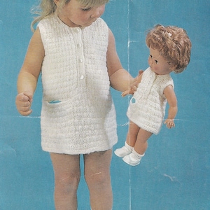 May include: A young girl in a white knitted dress holds a doll wearing a similar dress. Both dresses have a pocket on the front. The girl is wearing white socks and shoes.