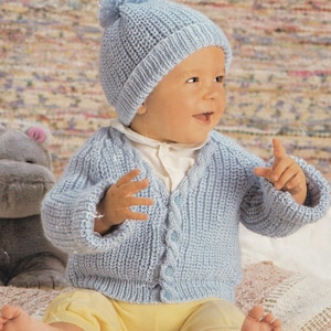 May include: A baby wearing a light blue knitted hat with a pom-pom and a matching cardigan. The cardigan has button closures. The baby is also wearing a white shirt and yellow pants.