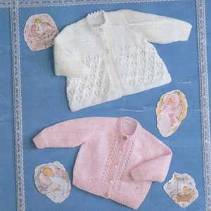 Puede incluir: Dos cárdigans de punto para bebé, uno blanco y otro rosa, con un delicado patrón de encaje. Ambos cárdigans tienen cierre de botones y cuello redondo.