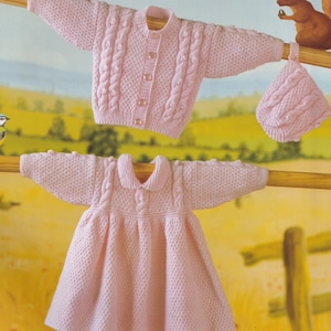 May include: A pink knitted baby dress and cardigan set with a matching hat. The set features a cable knit pattern and is hung on a wooden dowel.